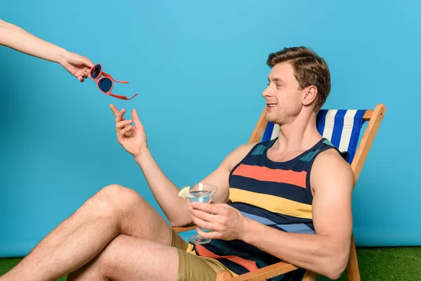 Частичный вид женщины, дающей солнцезащитные очки улыбающемуся мужчине, сидящему в шезлонге и держащему стакан коктейля на синем фоне — стоковое фото