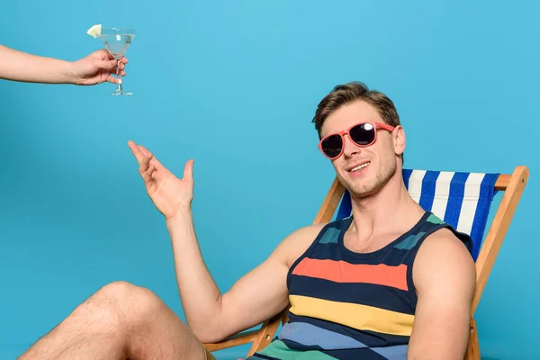 Vue recadrée de la femme donnant un verre de cocktail à l'homme en lunettes de soleil assis dans une chaise longue sur fond bleu — Photo de stock