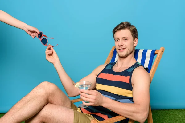 Vue recadrée de la femme donnant des lunettes de soleil à un homme souriant assis sur une chaise longue sur fond bleu — Photo de stock