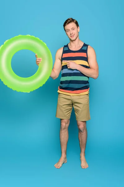 Lächelnder Mann in gestreiftem Singlet und kurzen Hosen, der mit dem Finger auf Schwimmring auf blauem Hintergrund zeigt — Stockfoto