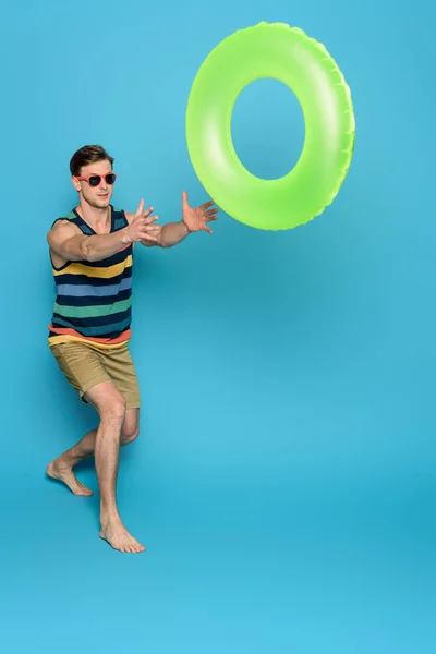 Jeune homme en singulet rayé et short jetant anneau gonflable sur fond bleu — Photo de stock