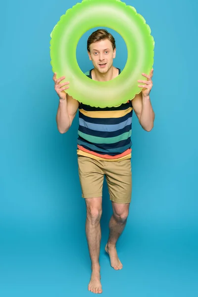 Positive young man in striped singlet and shorts holding swim ring and looking at camera on blue background — Stock Photo