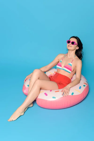 Happy woman in sunglasses sunbathing while sitting on inflatable ring on blue background — Stock Photo