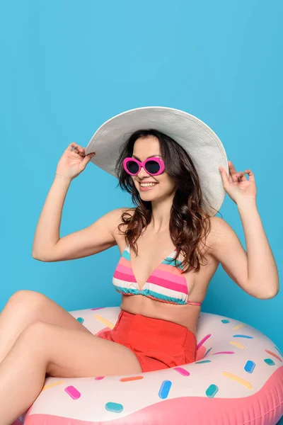 Mujer feliz y elegante en gafas de sol tocando sombrero de sol mientras está sentado en el anillo de natación sobre fondo azul - foto de stock