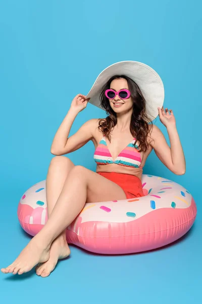 Cheerful, stylish woman in sunglasses touching sun hat while sitting on swim ring on blue background — Stock Photo