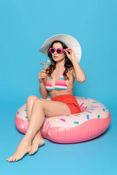 Femme élégante dans des lunettes de soleil et chapeau de soleil assis sur l'anneau de natation tout en tenant un verre de cocktail sur fond bleu — Photo de stock