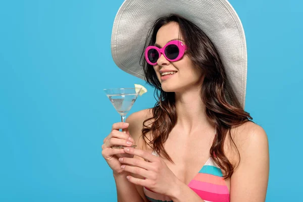 Mujer con estilo feliz en gafas de sol y sombrero de sol sosteniendo vaso de cóctel aislado en azul - foto de stock