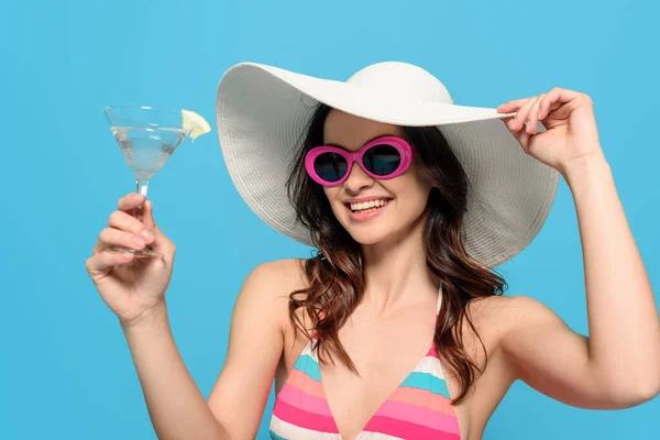 Femme élégante gaie dans des lunettes de soleil et chapeau de soleil tenant verre de cocktail isolé sur bleu — Photo de stock