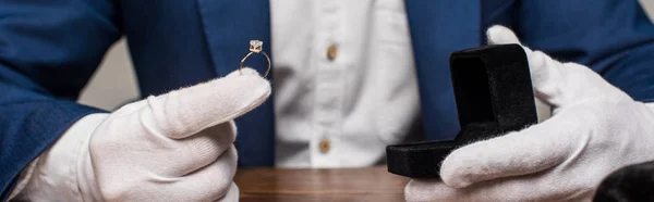 Cropped view of jewelry appraiser holding ring with gemstone and box on table isolated on grey, panoramic shot — Stock Photo