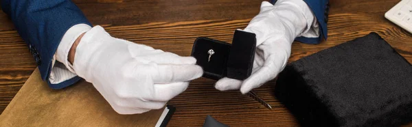 Cropped view of jewelry appraiser holding box with ring with gemstone near wooden table, panoramic shot — Stock Photo