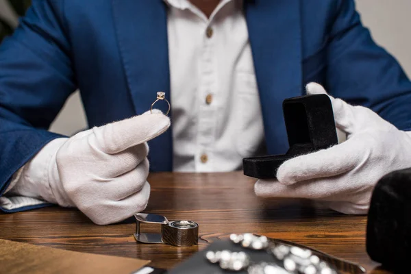 Обрезанный вид ювелира оценщик держа кольцо с драгоценным камнем и коробка на столе на сером фоне — стоковое фото