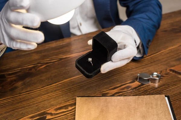 Vista recortada del anillo de sujeción del tasador de joyas con gemas en caja cerca de la lámpara en la mesa aislada en gris - foto de stock