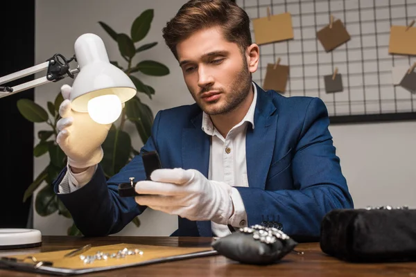 Handsome jewelry appraiser holding ring in box near lamp and gemstones and tools on table — Stock Photo