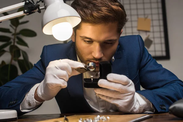 Bellissimo gioielliere valutatore esaminando anello con gemma in scatola vicino alla lampada sul tavolo — Foto stock