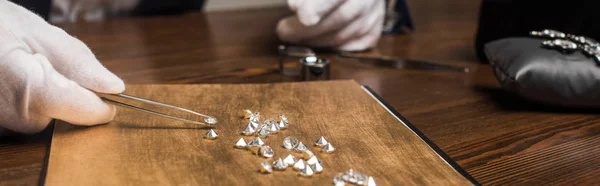 Vista recortada del tasador de joyas con pinzas que examinan las piedras preciosas en la mesa de madera aislada en gris, tiro panorámico - foto de stock