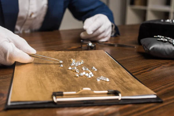 Vista ritagliata di perito di gioielli con pinze esaminando pietre preziose su tavola di legno in officina — Foto stock