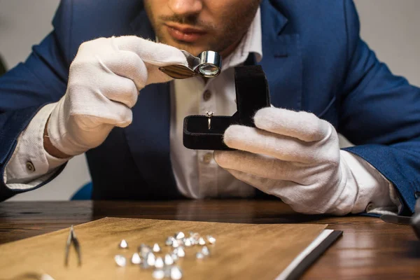 Vue recadrée de l'évaluateur de bijoux avec loupe examinant la pierre précieuse dans l'anneau à la table sur fond gris — Photo de stock