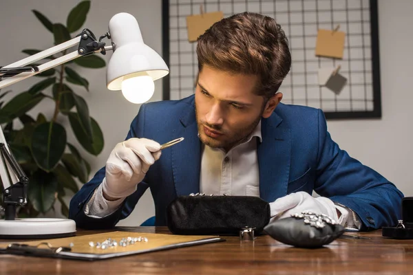 Handsome jewelry appraiser examining gemstone in pliers near jewelry on table in workshop — Stock Photo