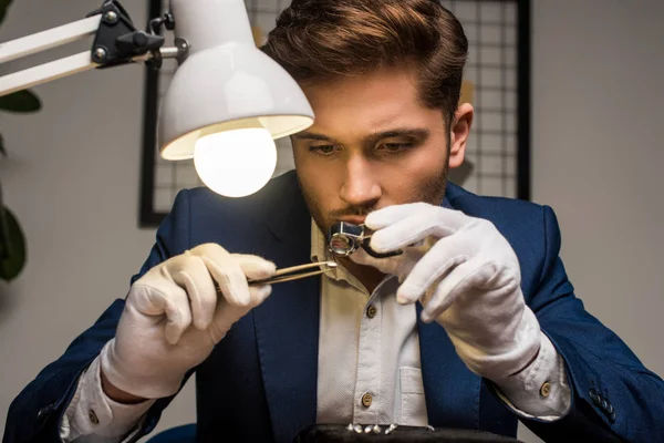 Handsome jewelry appraiser working with gemstone in tweezers and magnifying glass at table in workshop — Stock Photo