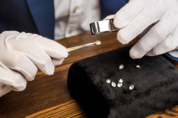 Vue rapprochée de l'évaluateur de bijoux examinant la pierre précieuse dans une pince à épiler avec loupe à table — Photo de stock