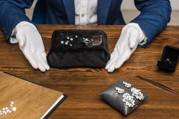 Vue recadrée de l'évaluateur de bijoux dans des gants près de pierres précieuses près de bijoux sur une table en bois — Photo de stock