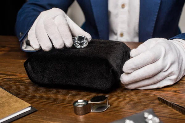 Vue recadrée de l'évaluateur de bijoux dans des gants tenant la pierre précieuse près des outils sur la table sur fond gris — Photo de stock