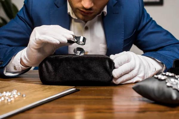 Vista recortada del tasador de joyas examinando piedras preciosas cerca de pendientes en la almohada de joyería en la mesa - foto de stock