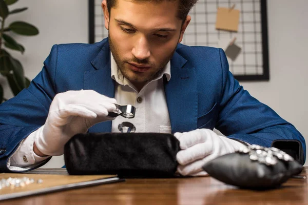 Schöner Schmuckgutachter hält Vergrößerungsglas in der Hand, während er Edelstein in der Nähe von Ohrringen auf dem Tisch untersucht — Stockfoto