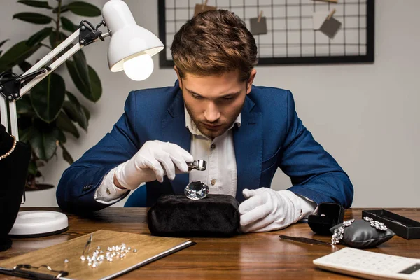 Jewelry appraiser with magnifying glass examining gemstone near jewelry on table in workshop — Stock Photo