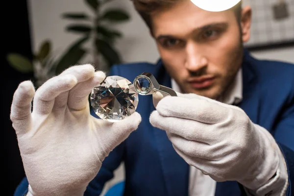 Focus selettivo del perito di gioielli in guanti esaminando la gemma con lente d'ingrandimento in officina — Foto stock