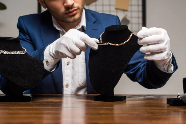 Vista recortada del tasador de joyas en guantes con pinzas cerca del collar en el soporte del collar en la mesa - foto de stock