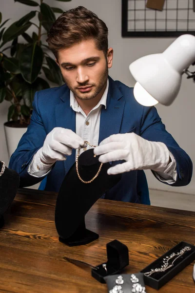 Schöner Schmuckgutachter in Handschuhen mit Halskette in der Nähe von Halskettenständer und Schmuck auf dem Tisch — Stockfoto