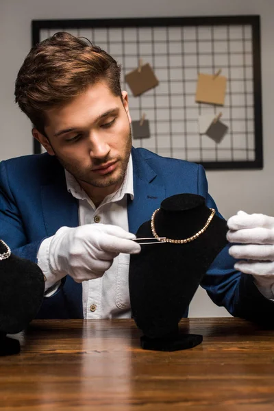 Apprezzatore di gioielli bello tenendo pinzette vicino collana sul piedistallo sul tavolo in officina — Foto stock