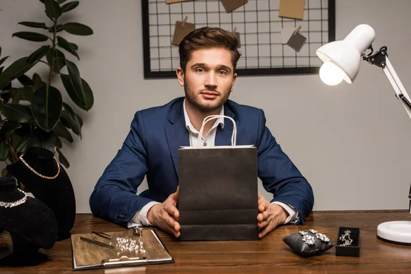 Schöner Schmuckgutachter blickt in Kamera, während er Papiertüte in der Nähe von Schmuck auf Tisch in Werkstatt hält — Stockfoto