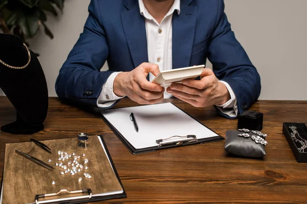 Vue recadrée de l'évaluateur de bijoux en utilisant une calculatrice près du presse-papiers et des bijoux sur la table — Photo de stock