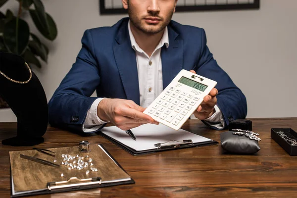 Vue recadrée de l'évaluateur de bijoux montrant la calculatrice près du presse-papiers et des bijoux sur la table — Photo de stock