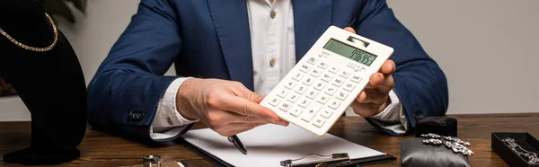 Vue recadrée de l'évaluateur de bijoux montrant la calculatrice près du presse-papiers et des bijoux sur la table, vue panoramique — Photo de stock