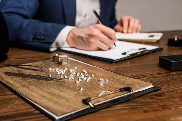 Selective focus of gemstones and tools near jewelry appraiser writing on clipboard on table isolated on grey — Stock Photo