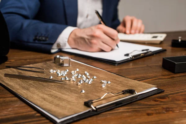 Concentration sélective des pierres précieuses et des outils près de l'évaluateur de bijoux écrivant sur le presse-papiers sur la table isolé sur gris — Photo de stock