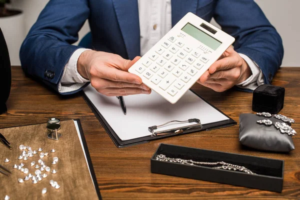 Vue recadrée de l'évaluateur de bijoux tenant la calculatrice près du presse-papiers et des bijoux avec des pierres précieuses sur la table — Photo de stock