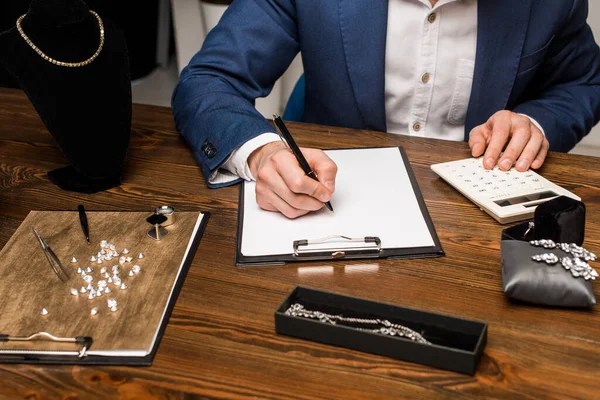 Ausgeschnittene Ansicht des Schmuckgutachters mit Taschenrechner und Schreiben auf Klemmbrett in der Nähe von Schmuck auf Holztisch — Stockfoto