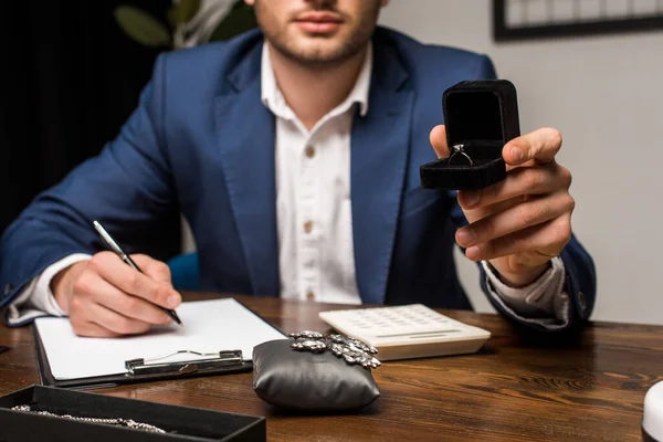 Cropped view of jewelry appraiser holding box with ring and writing on clipboard near jewelry on wooden table — Stock Photo