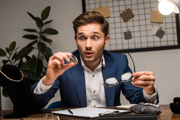 Vorhandener Schmuckgutachter hält Edelstein und Brille neben Schmuck auf Tisch in Werkstatt — Stockfoto