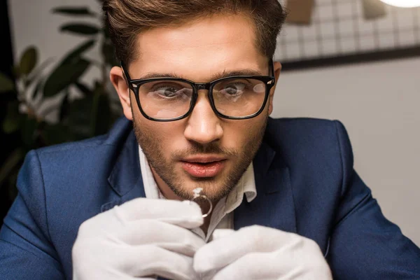 Shocked jewelry appraiser in gloves holding jewelry ring with gemstone in workshop — Stock Photo