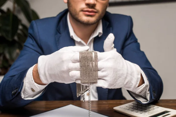 Cropped view of jewelry appraiser in gloves holding necklace near calculator and clipboard on table in workshop — Stock Photo