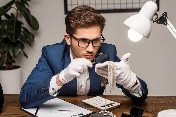 Evaluador de joyas examinando collar con lupa cerca de la calculadora, portapapeles y joyas en la mesa - foto de stock