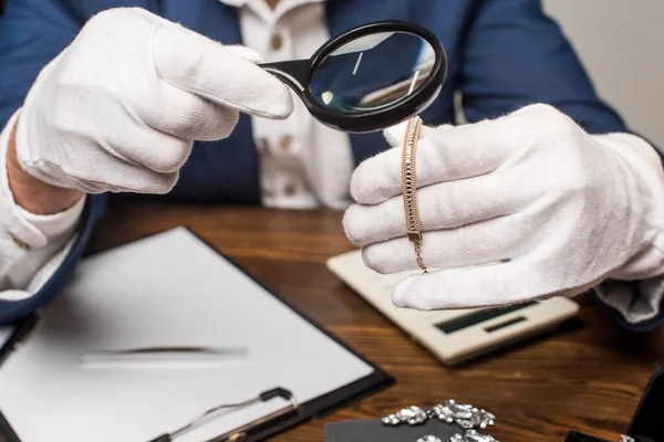 Ausgeschnittene Ansicht des Schmuckgutachters untersucht Halskette mit Lupe in der Nähe von Taschenrechner, Klemmbrett und Ergebnis auf Tisch isoliert auf grau — Stockfoto
