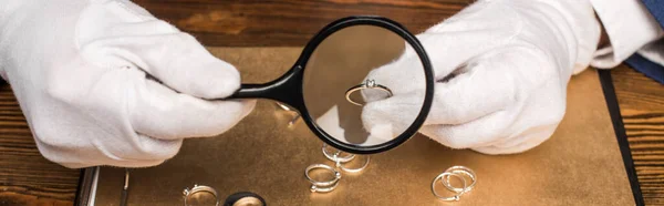 Cropped view of jewelry appraiser holding jewelry ring and magnifying glass near board on table, panoramic shot — Stock Photo