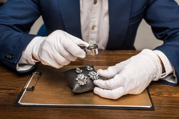 Vue recadrée de l'évaluateur de bijoux tenant loupe et gains à bord sur table isolé sur gris — Photo de stock