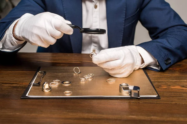 Vista recortada del tasador de joyas con lupa examinando el anillo de joyería cerca de la tabla sobre fondo gris - foto de stock